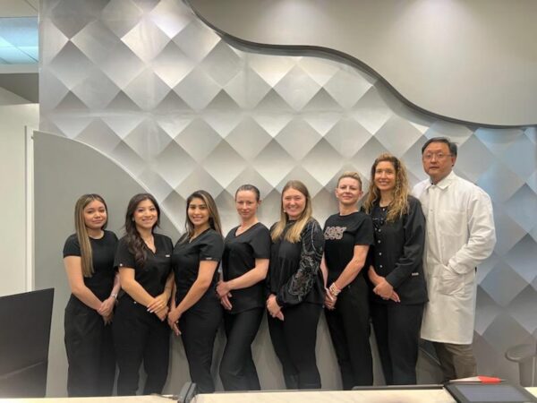 Group of seven contrast-clad professionals posing for a photo in a modern clinic reception; includes six women in black scrubs and a man in a white lab coat.