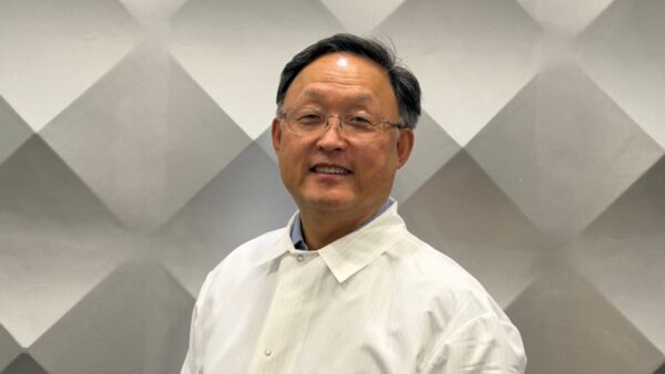 Smiling man with glasses in a light striped shirt, standing in front of a gray diamond-pattern wall.